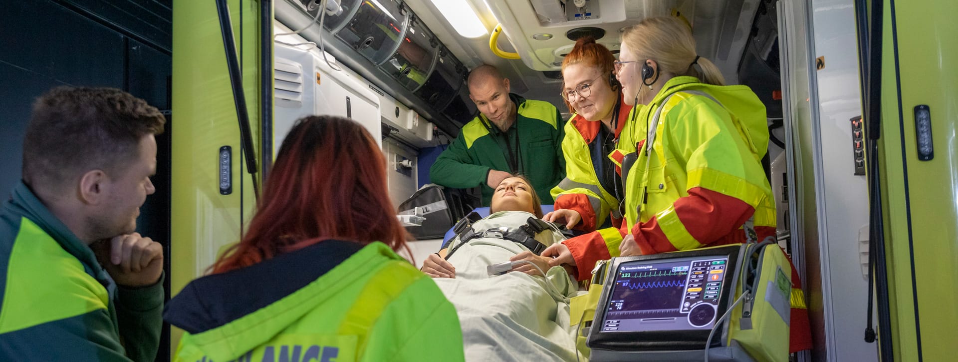 Kaksi miestä ja kolme naista harjoittelemassa ambulanssisimaattorin sisällä hoitotoimenpidettä.