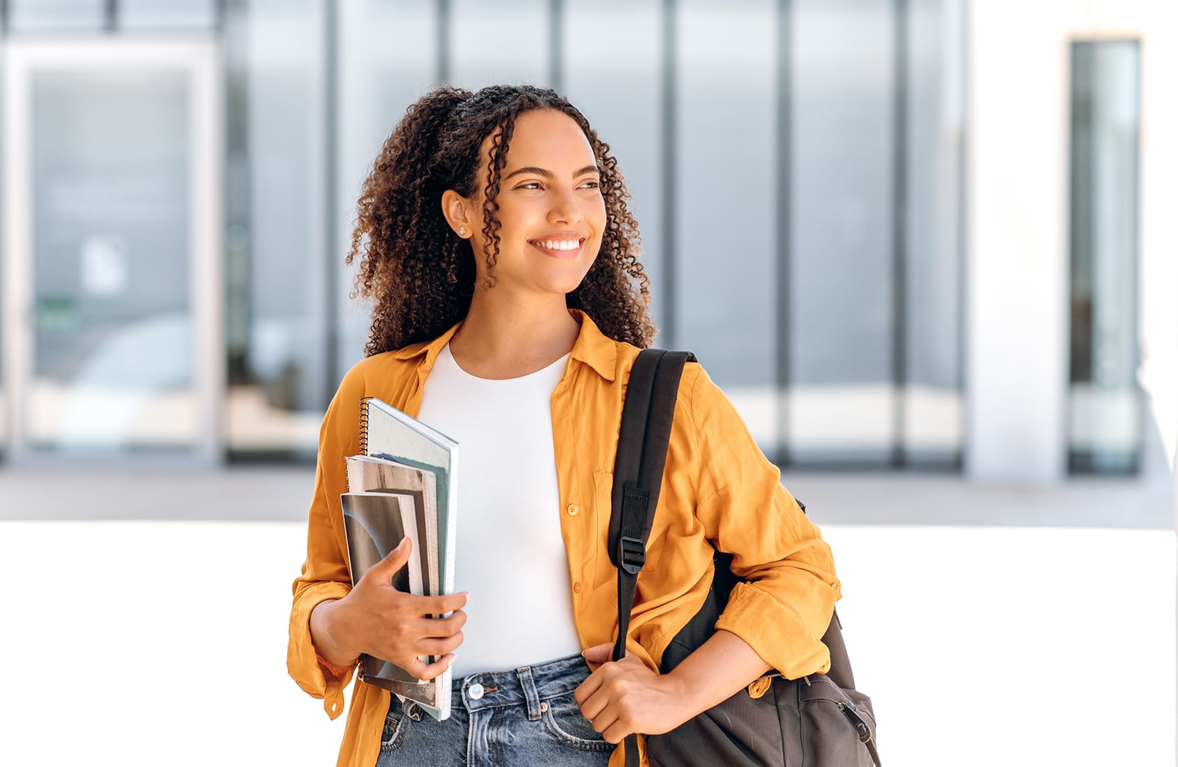 Happy female student. Positive female student, brazilian or hispanic nationality, with a backpack, holding books and notebooks in her hand, stand near the university campus, looks away and smiling