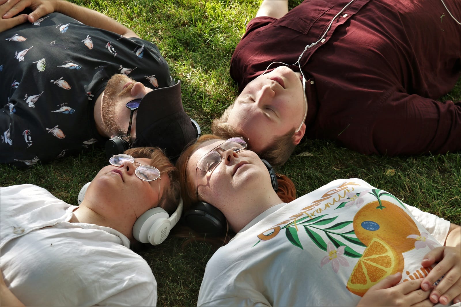 Valokuva. Nuoria henkilöitä makaamassa nurmikolla päätä vastakkain. Neljä henkilöä.