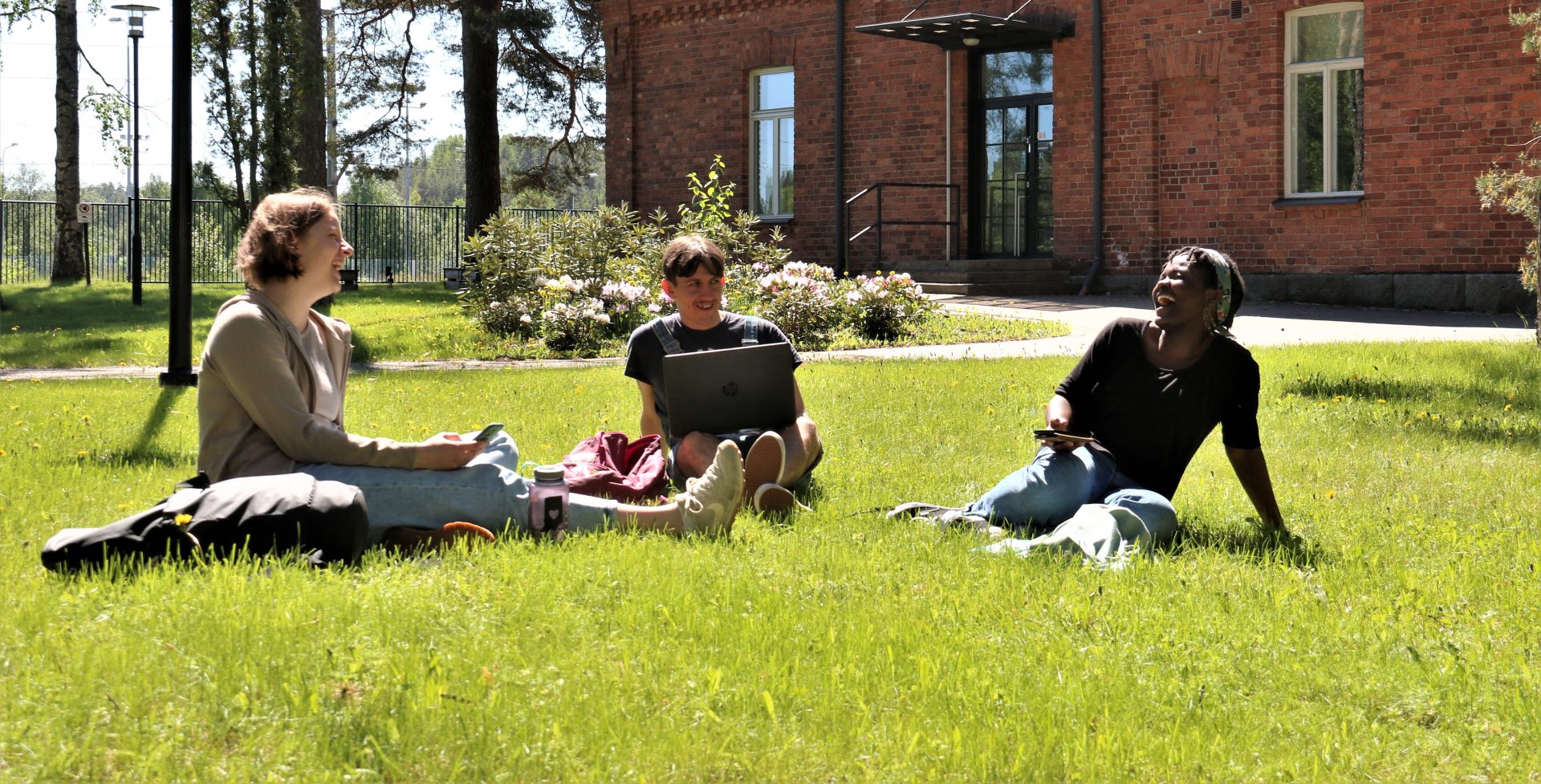 Valokuva. Kolme henkilöä istumassa nurmikolla. Aurinkoinen sää.