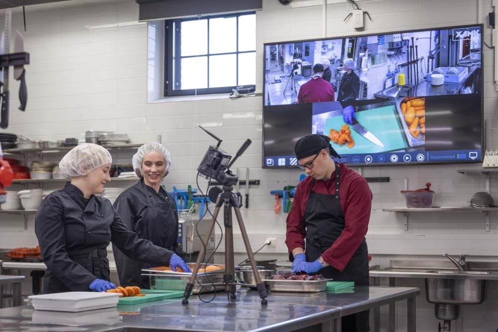 Kolme opiskelijaa työskentelee RestoLab-virtuaalikoulutustilan keittiössä. Taustalla näytöllä videokuvaa heidän työskentelystään.