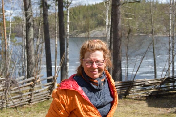 Opas-Tiina yrittäjä, Tiina Leinonen poseeraa metsäisessä maisemassa. Taustalla vettä, joko joki tai järvi.