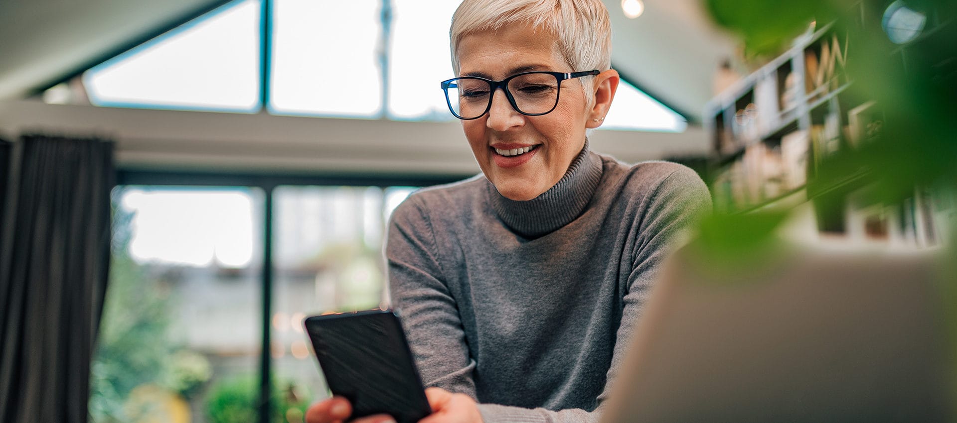 Close-up portrait of a happy senior woman looking at smart phone while sitting at desk with laptop.