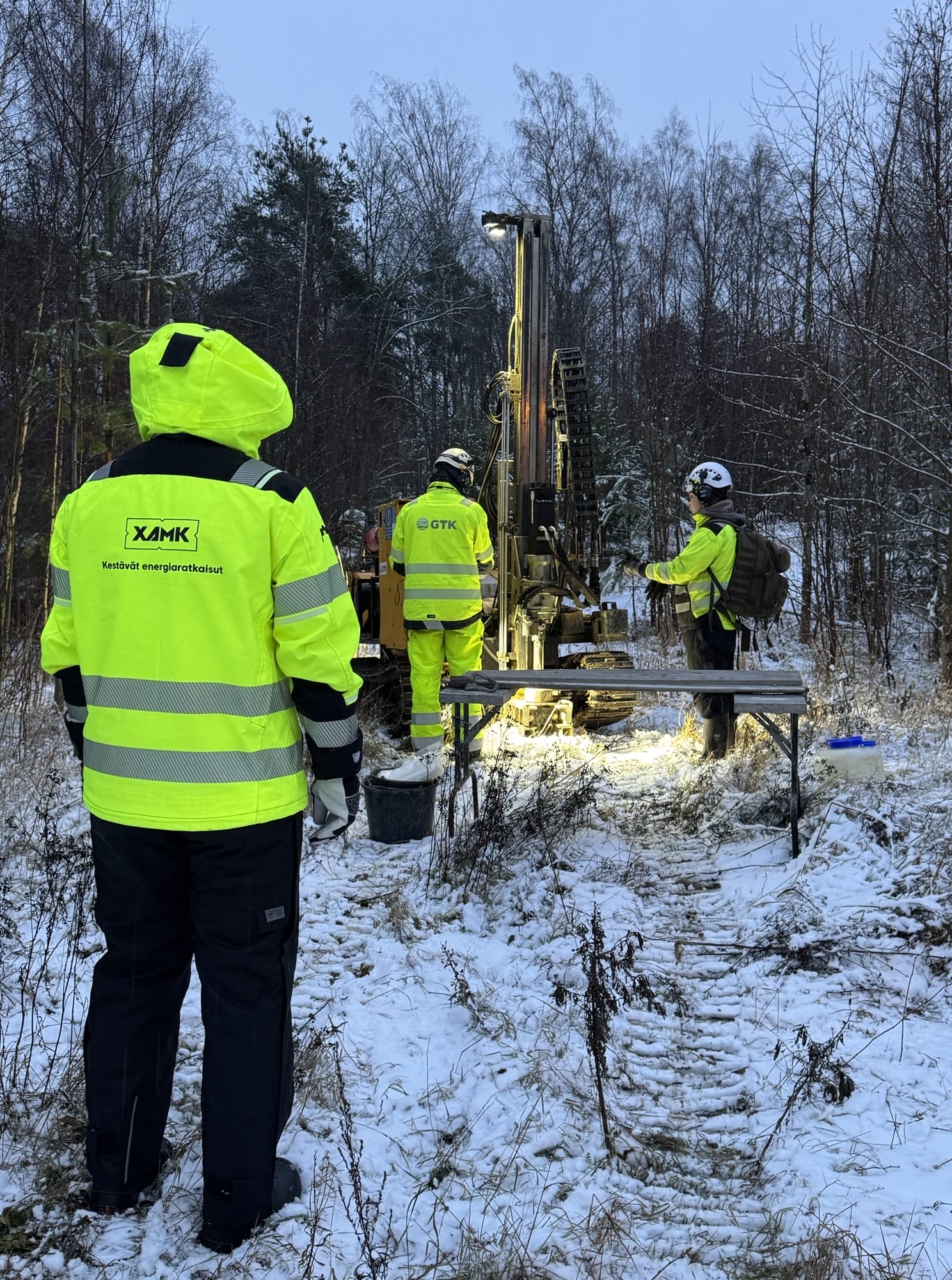 Metsämaastossa suuri pora ja työvaatteisiin pukeutuneita ihmisiä.